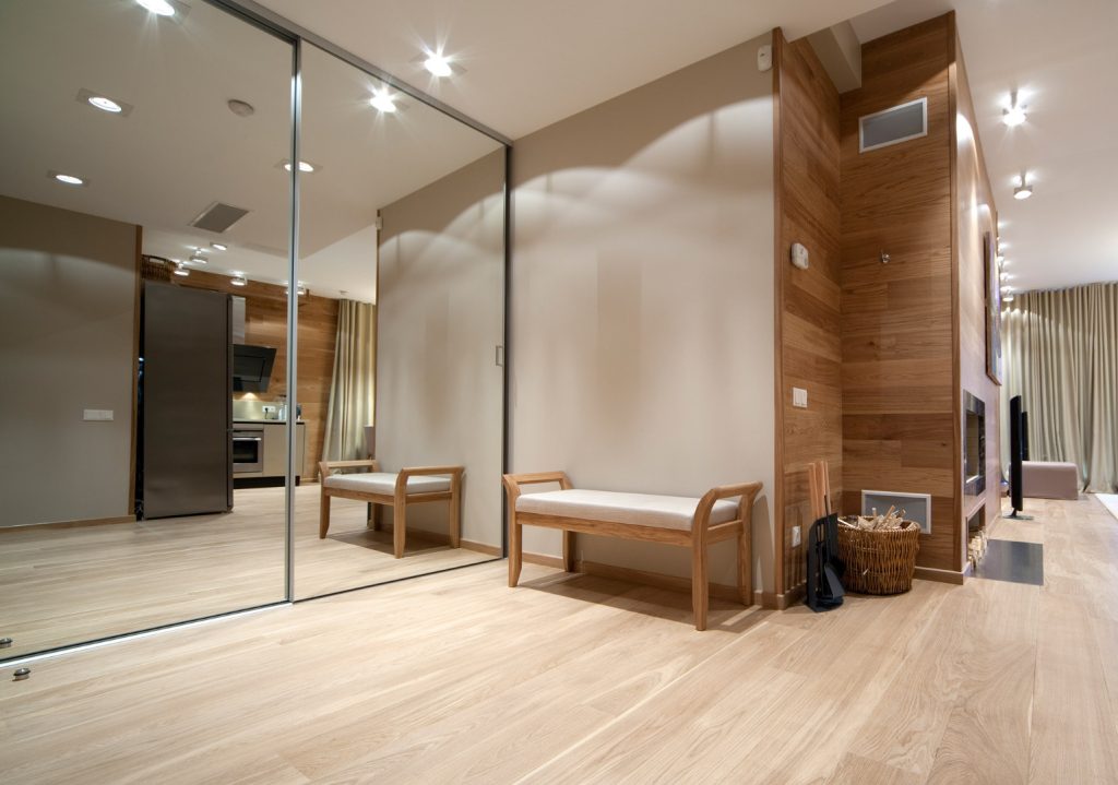 Een moderne woonkamer met houten vloeren, een houten bankje met witte kussens, een spiegelwand en een open keuken op de achtergrond.