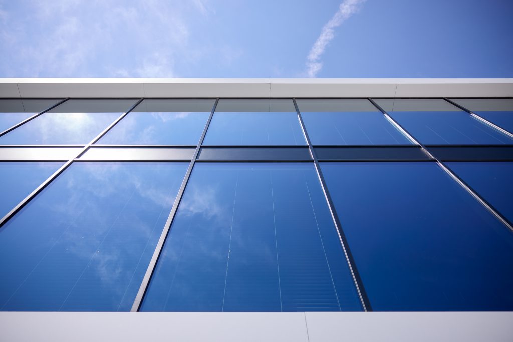 Moderne gevel met grote reflecterende glazen ramen en weerspiegeling van blauwe lucht met wolken.